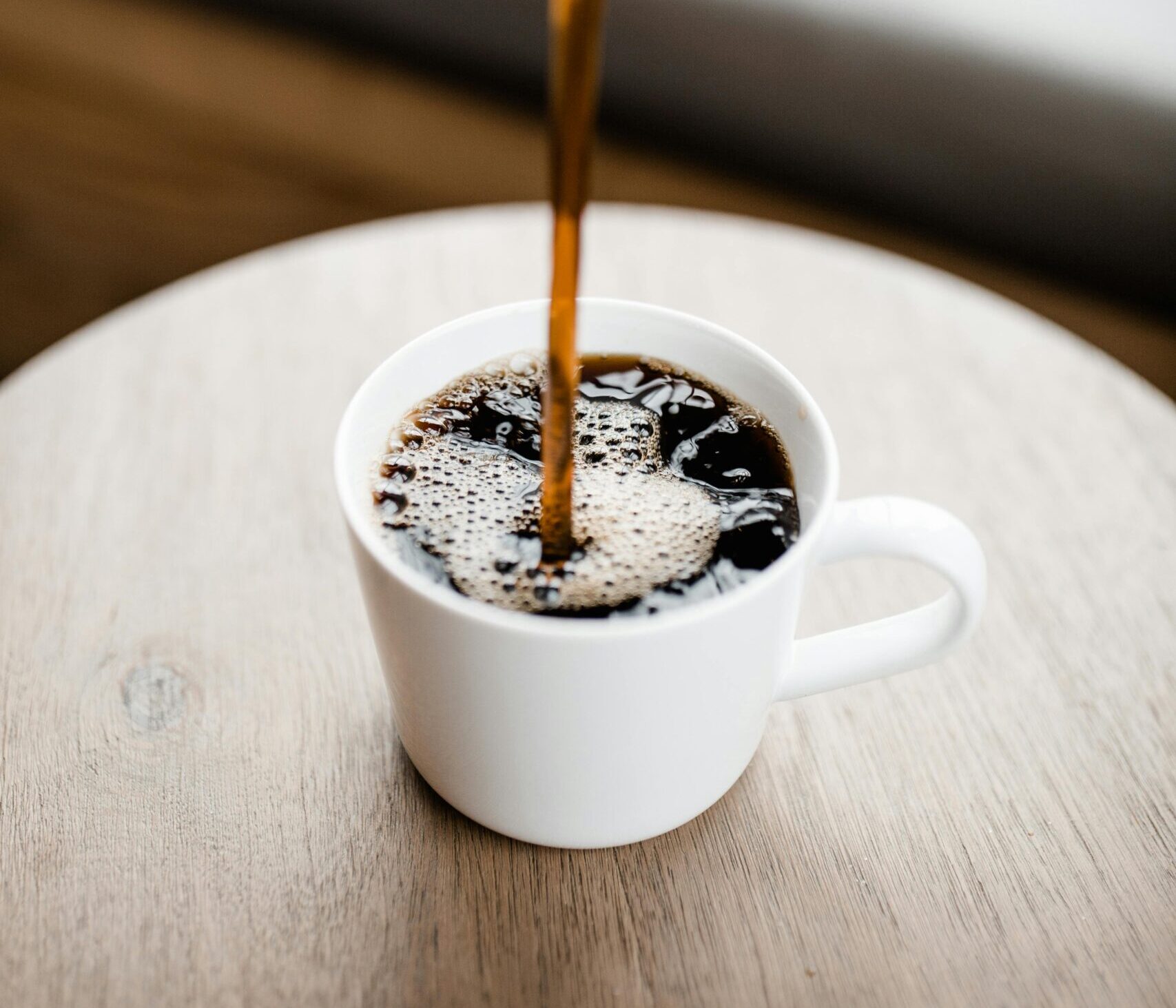 Steaming black coffee being poured into a white ceramic mug on a wooden table.